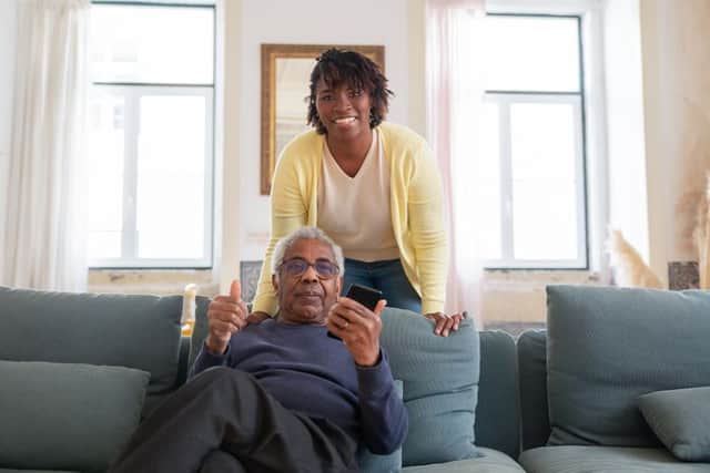 An elderly man giving a thumbs up from the couch while his caregiver smiles behind him.