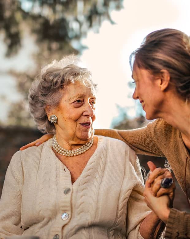 An elderly woman in pearls holding hands with her caregiver in a golden garden, smiling warmly at each other.