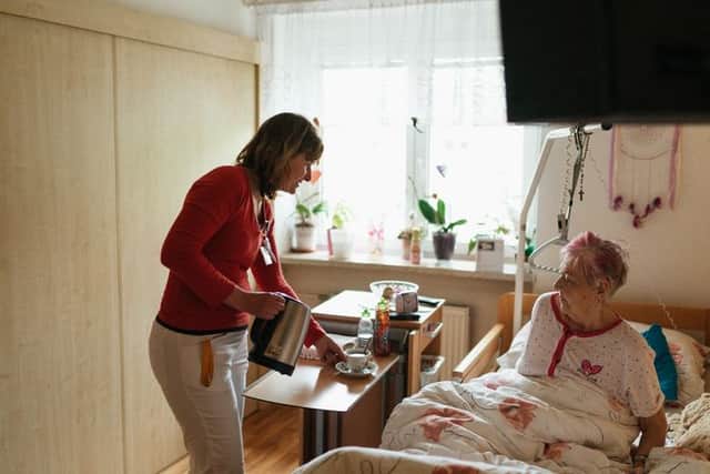 A caregiver pouring tea for a smiling elderly woman in her sunny bedroom.