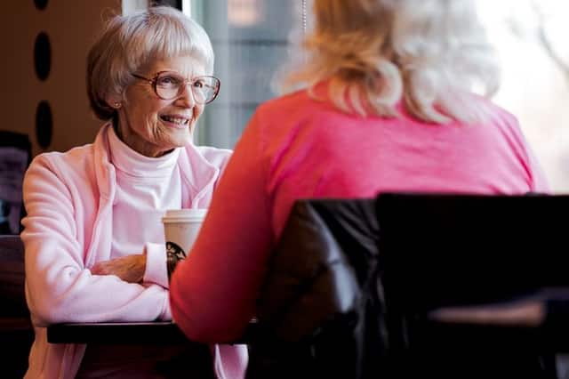A senior woman in pink smiling over coffee with a friend by a sunny window.